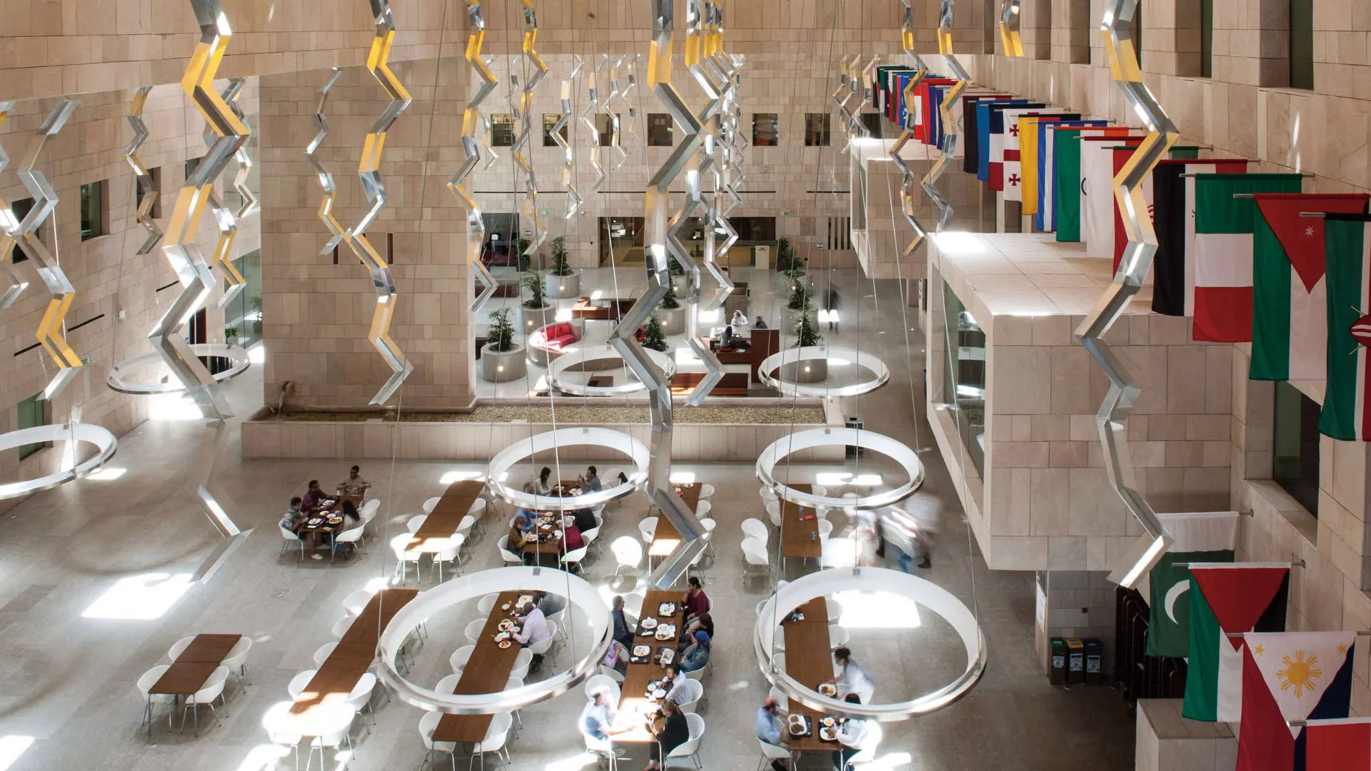Inside of a light colored spacious building. Flags hang on the right side wall. Tables at the bottom floor where people engage in conversations.