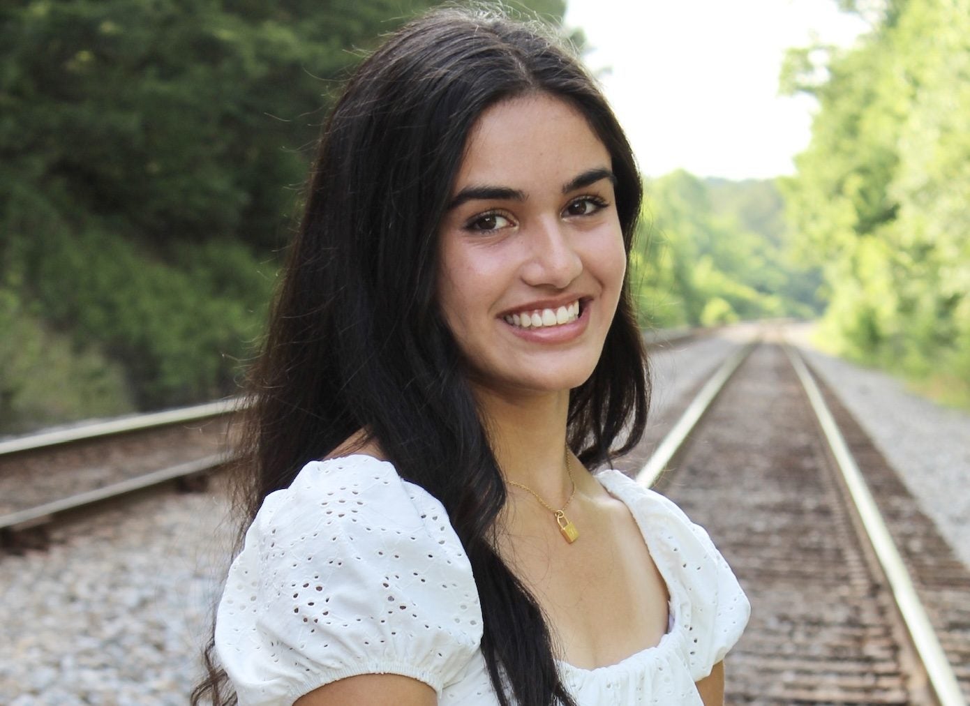 A young women with long dark hair wearing a white broderie top and standing on railroad tracks.