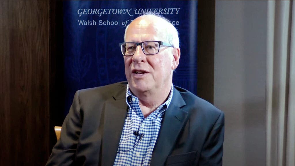 Richard Hluchan speaking while sitting down. Behind him a banner for the Georgetown University School of Foreign Service