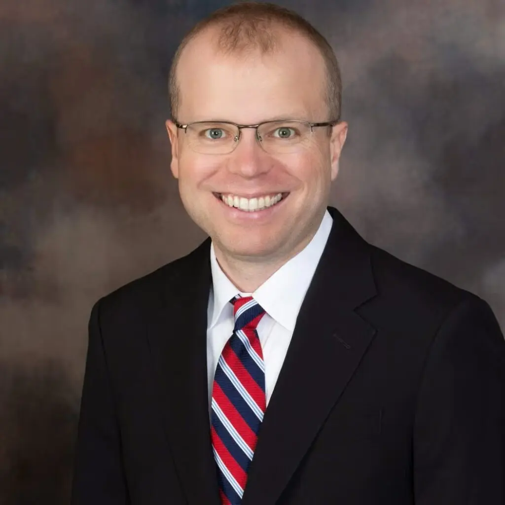 Robert Lyons wearing a black blazer, a red and navy striped tie and a white button-down shirt and glasses.