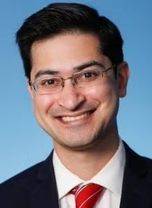 Portrait of a smiling individual wearing glasses, a white shirt, and a red tie with blue stripes, set against a solid blue background.