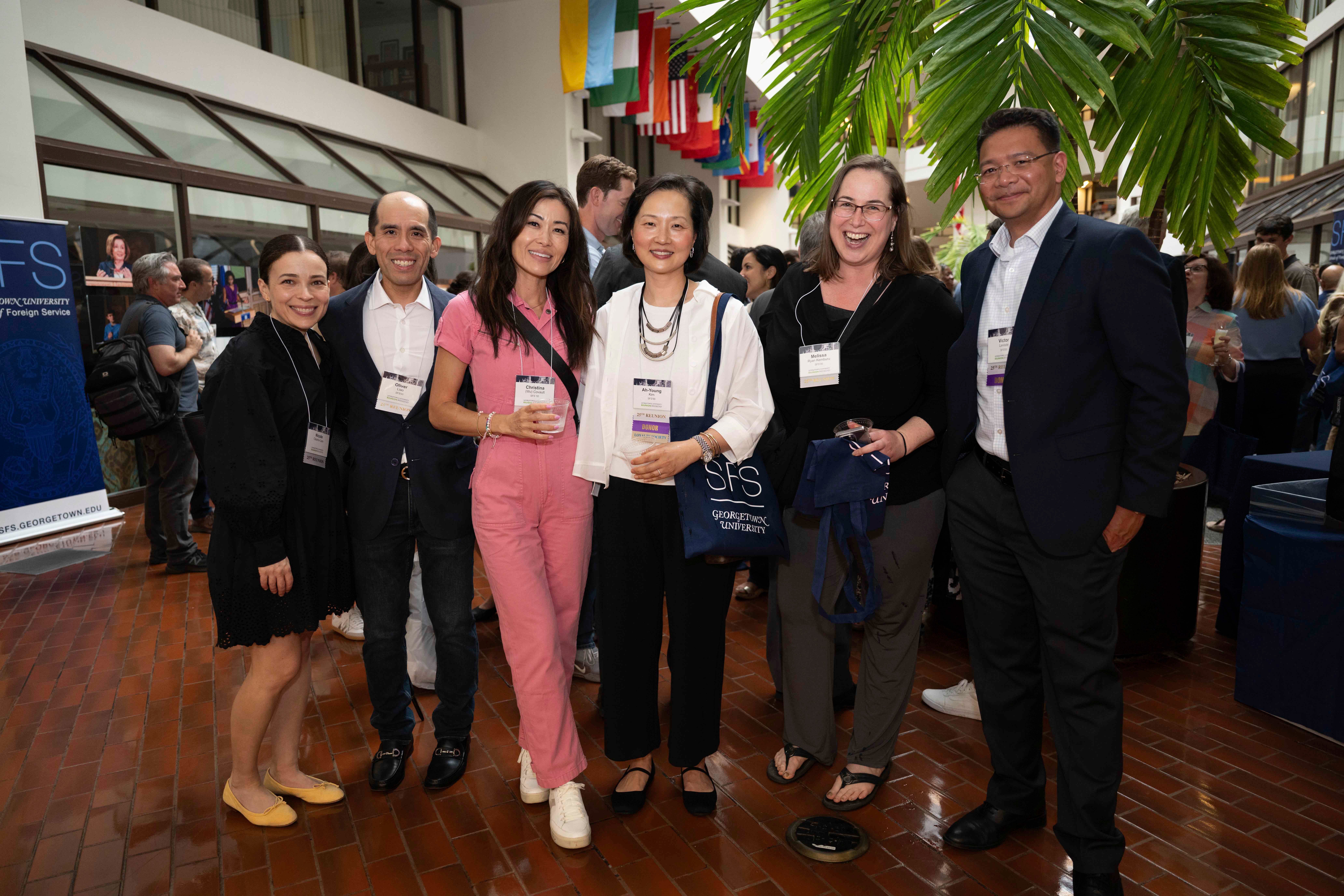 Group of six professionals networking at a conference, smiling and holding drinks, with banners and colorful flags in the background.