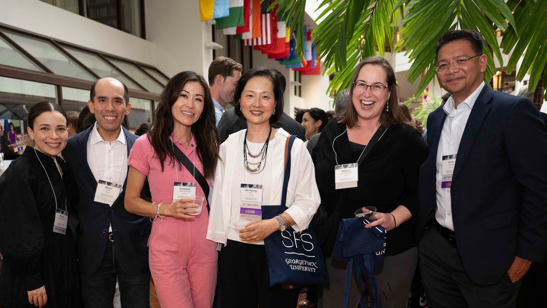 Group of six professionals networking at a conference, smiling and holding drinks, with banners and colorful flags in the background.
