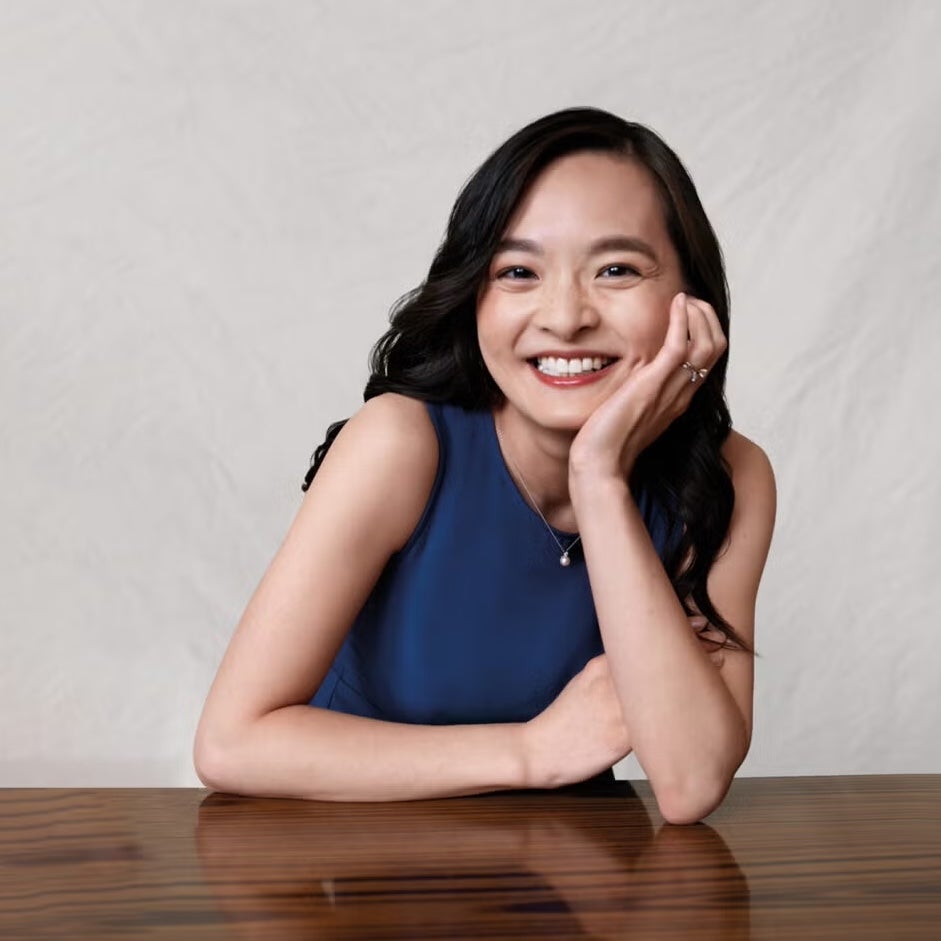 Sonia Su wearing a blue shirt and leaning on a table while smiling.