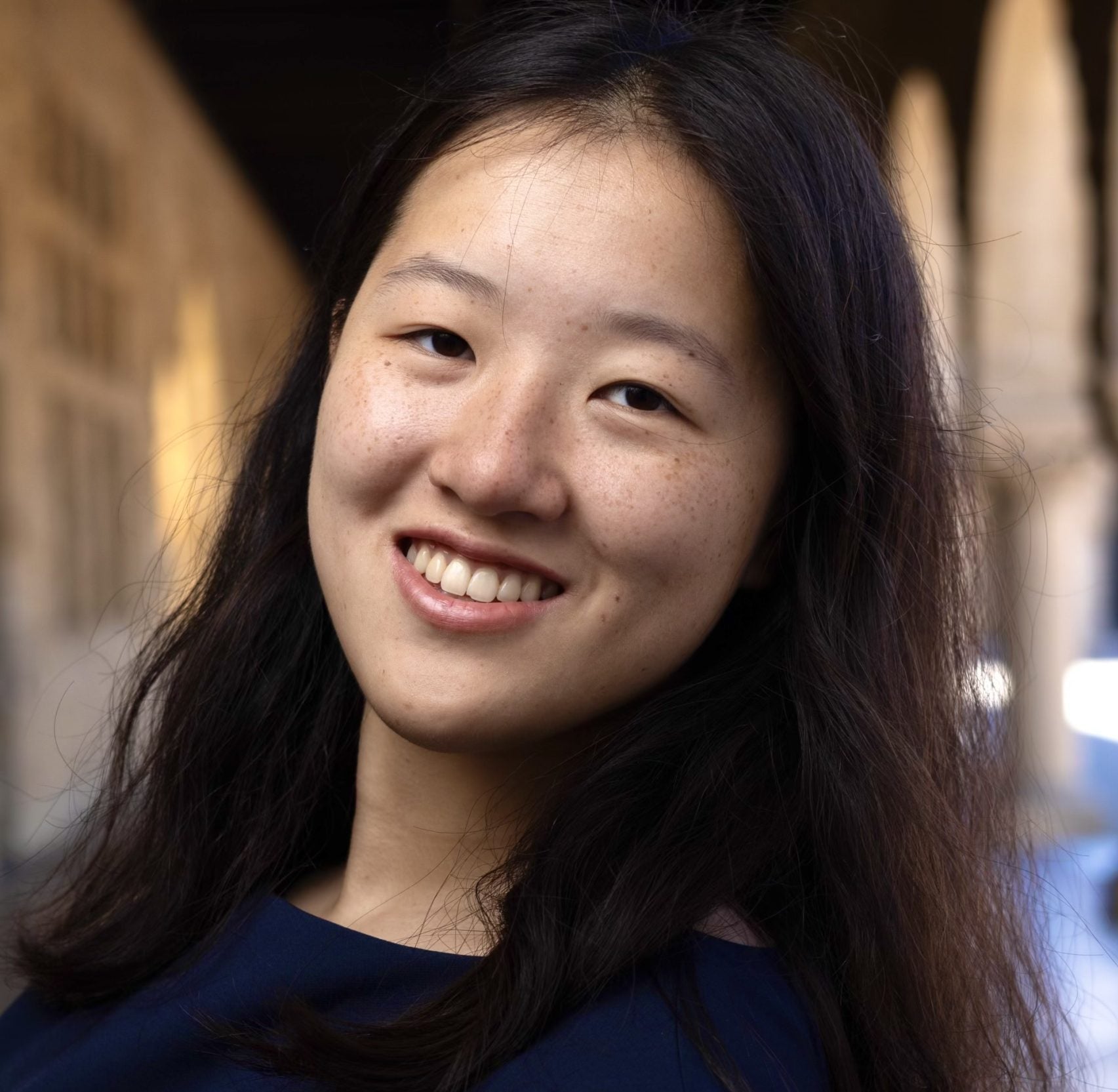 A young woman with dark brown hair wearing a dark blue shirt and smiling.