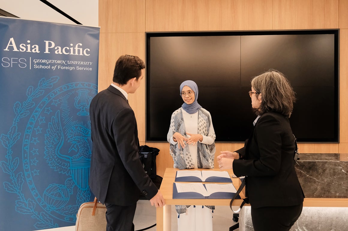 Three people engaging in conversation while standing around a table. The table has folders with papers. Behind them a banner for Georgetown SFS Asia Pacific