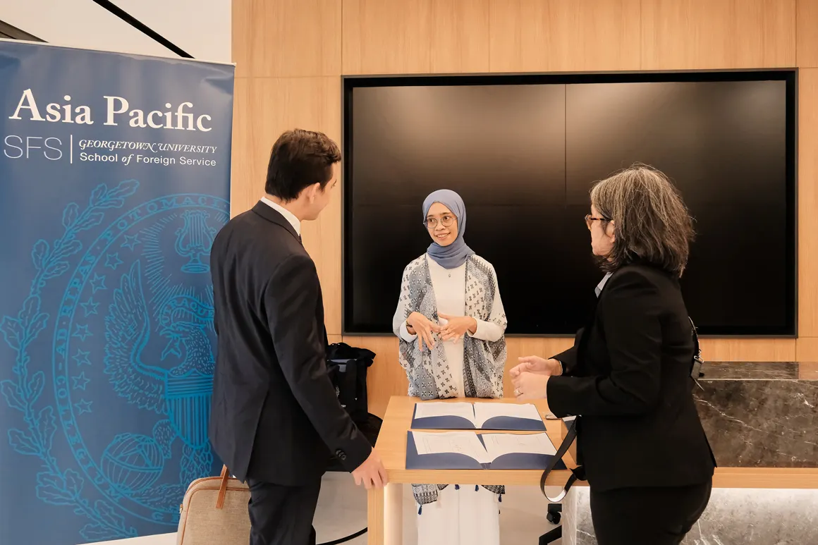 Three people engaging in conversation while standing around a table. The table has folders with papers. Behind them a banner for Georgetown SFS Asia Pacific