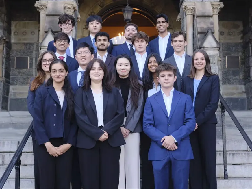 A group of 16 students wearing business attire standing in front of building steps smiling.