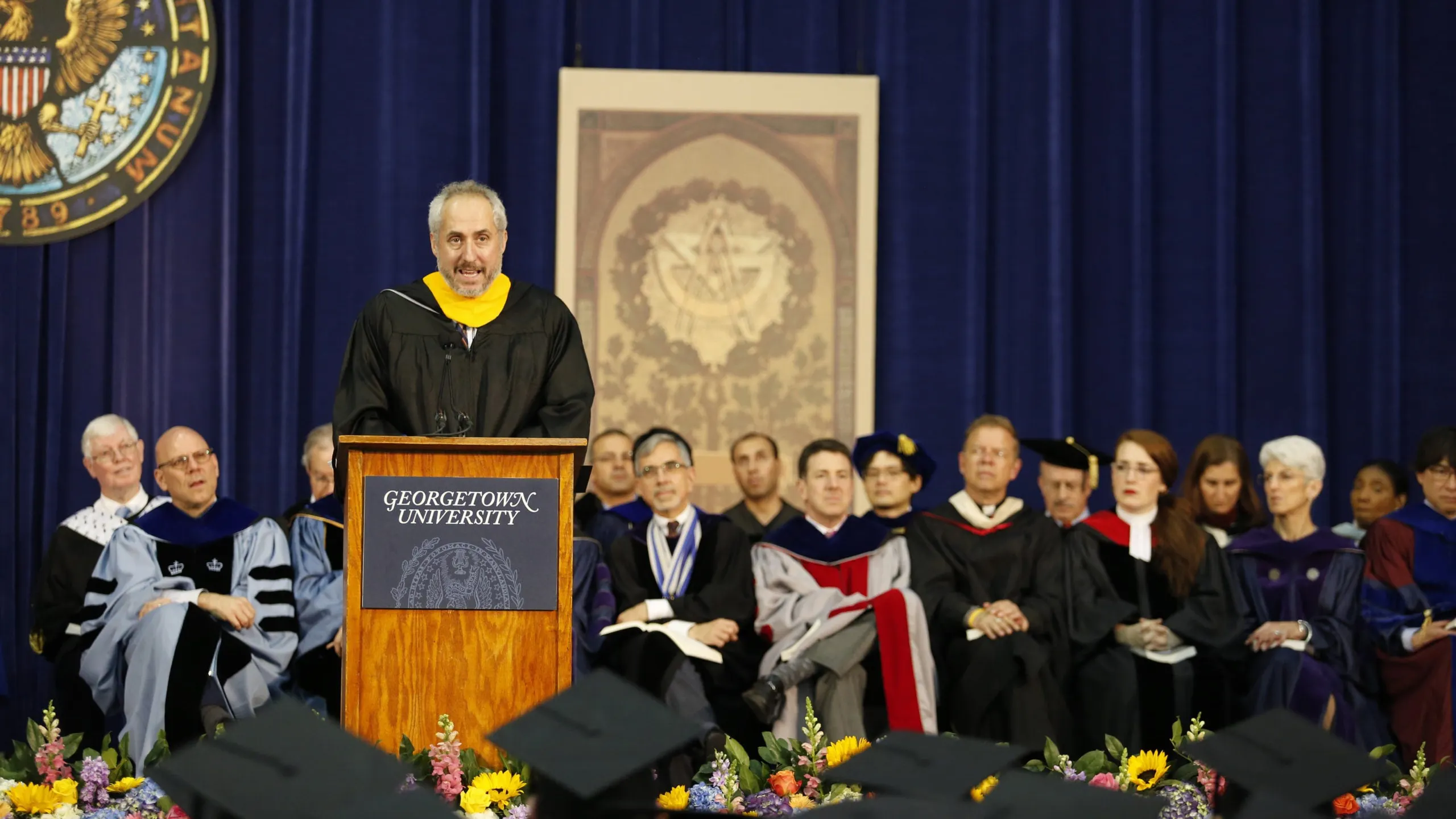 Stephane Dujarric in commencement regalia delivering a speech on a Georgetown University podium. Behind him, men and women in commencement gowns.