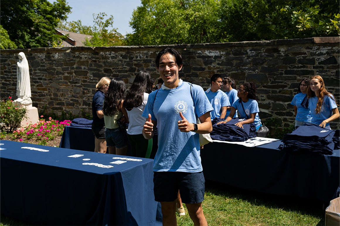 Student in the lawn wearing a blue Georgetown shirt with thumbs up. Behind him students gathered next to a table picking u shirts.