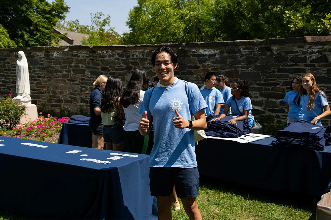 Student in the lawn wearing a blue Georgetown shirt with thumbs up. Behind him students gathered next to a table picking u shirts.