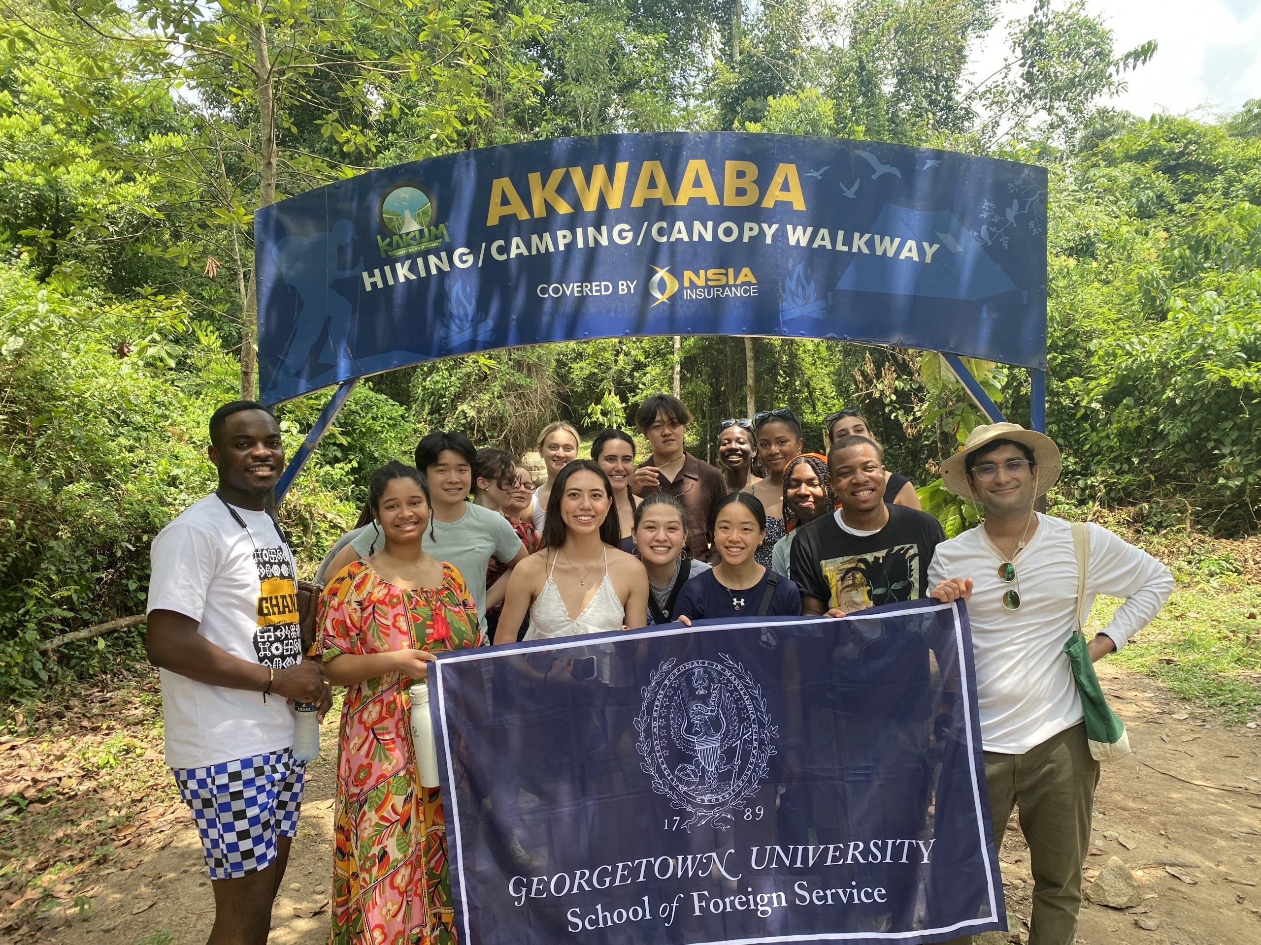Large group of students wearing summer hiking clothes hold Georgetown School of Foreign Service banner in front of entrance to Akwaaba walkway.