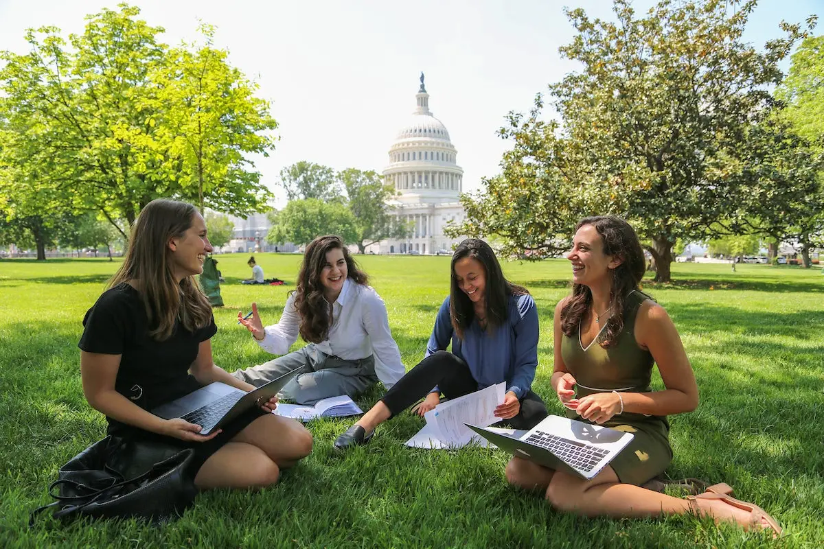 For young women in business attire sitting in the grass in front of the Capitol building, talking to each other, with laptops, smiling.