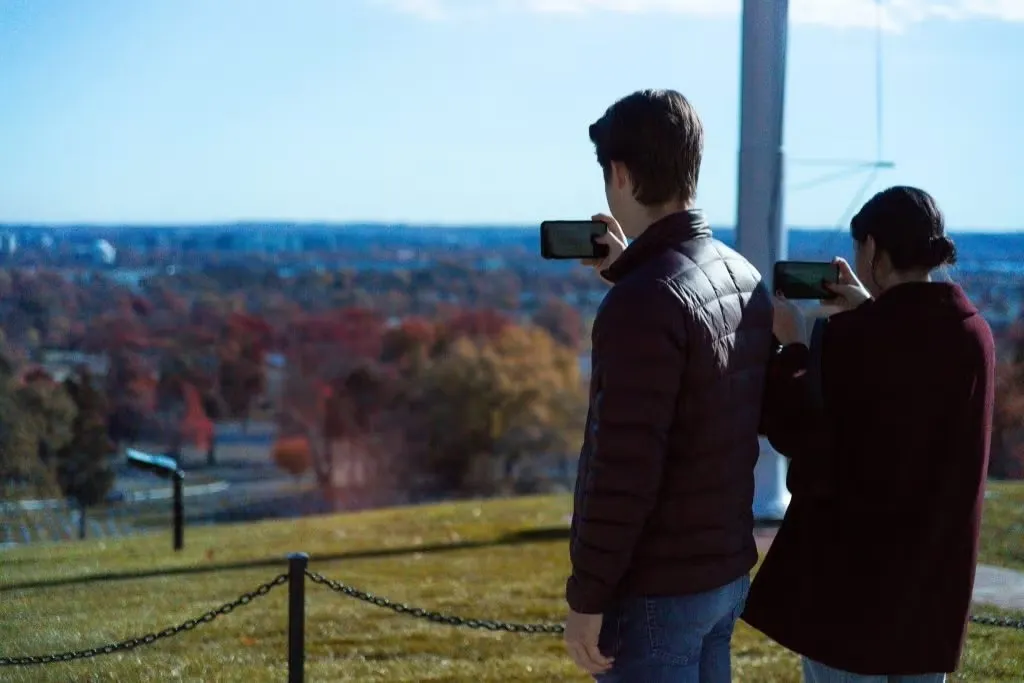 Two people taking photos with their smartphones overlooking a scenic landscape from a high vantage point.