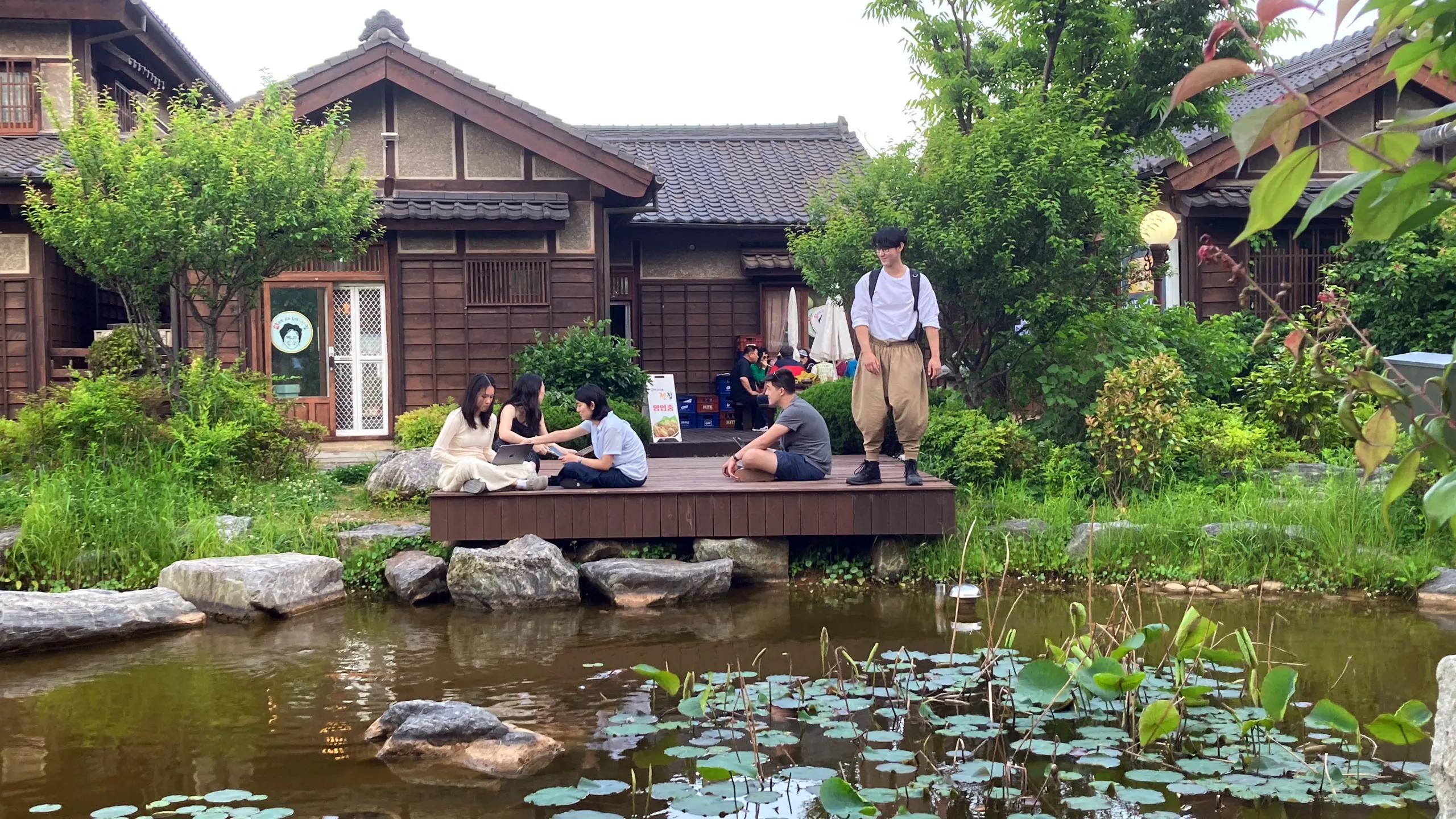 Five students in Japan sitting on a deck by a pond. Behind them, houses.