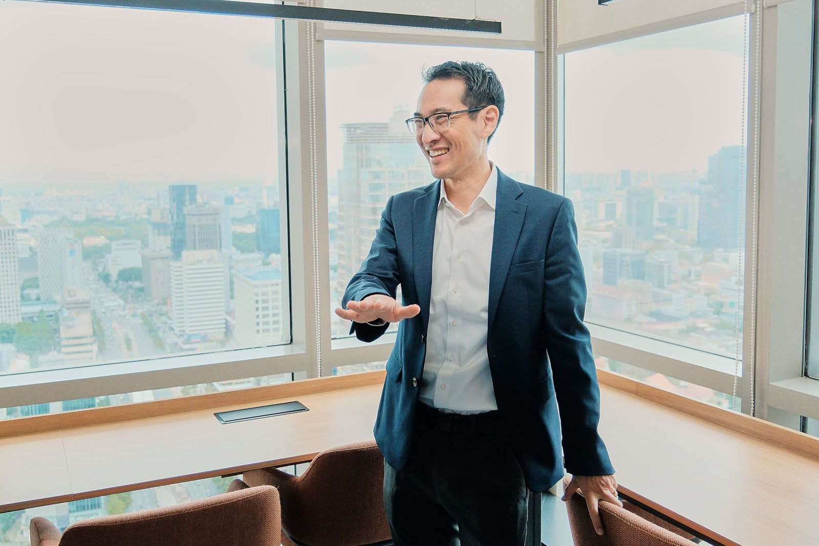 An Asian man in a blue suit stand in front of a window overlooking a city skyline.