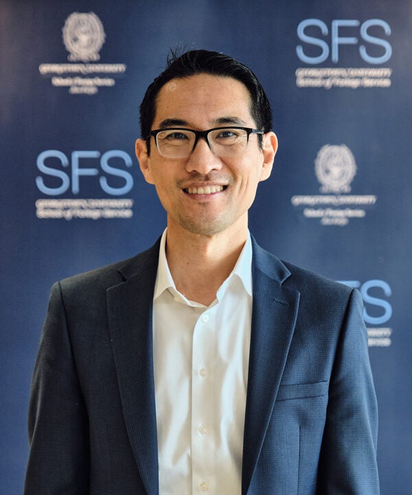 Headshot of Yuhki Tajima, smiling, wearing a suit. Behind him is a Georgetown SFS Asia Pacific backdrop.