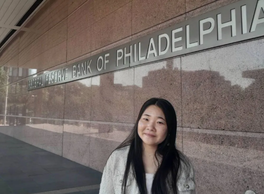A young woman with long, brown hair wearing a white jacket and standing in front of a building that says Federal Reserve Bank of Philadelphia