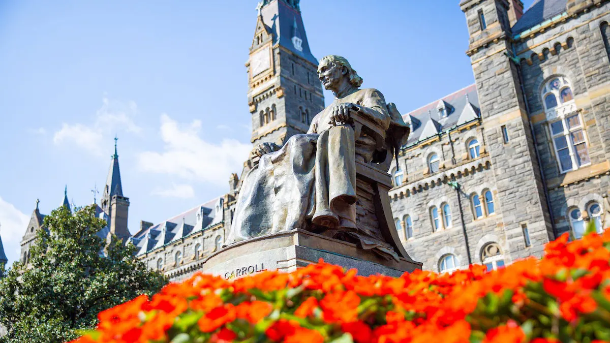 Statue of John Carroll sitting on a chair. Underneath him, there is a bed of red flowers. Behind him the Healy building.