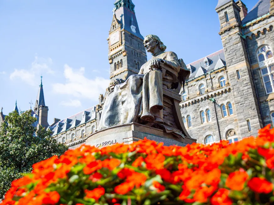 Statue of John Carroll sitting on a chair. Underneath him, there is a bed of red flowers. Behind him the Healy building.