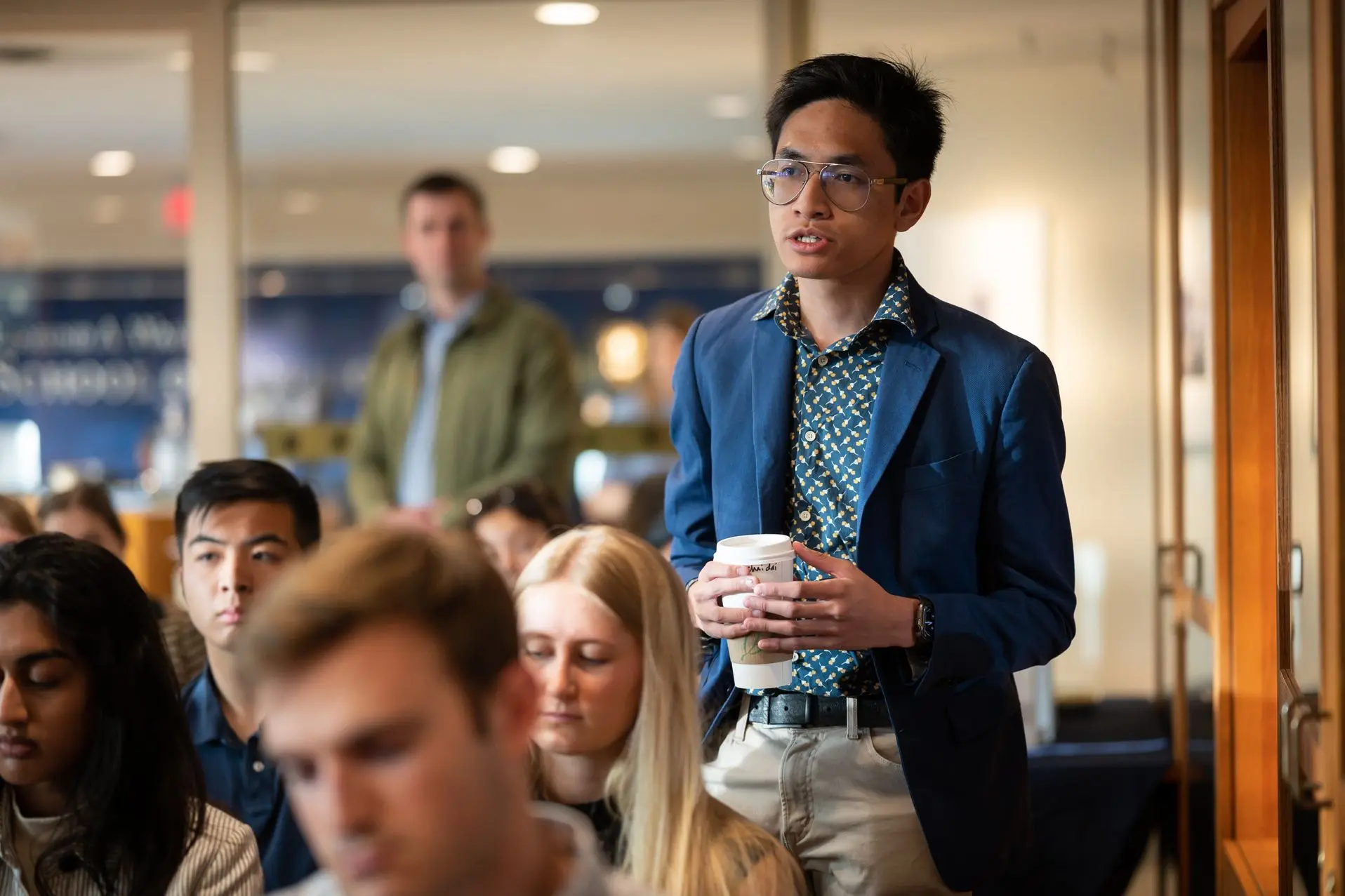 A student wearing a navy blazer and khakis asks a question during a talk while other students listen.