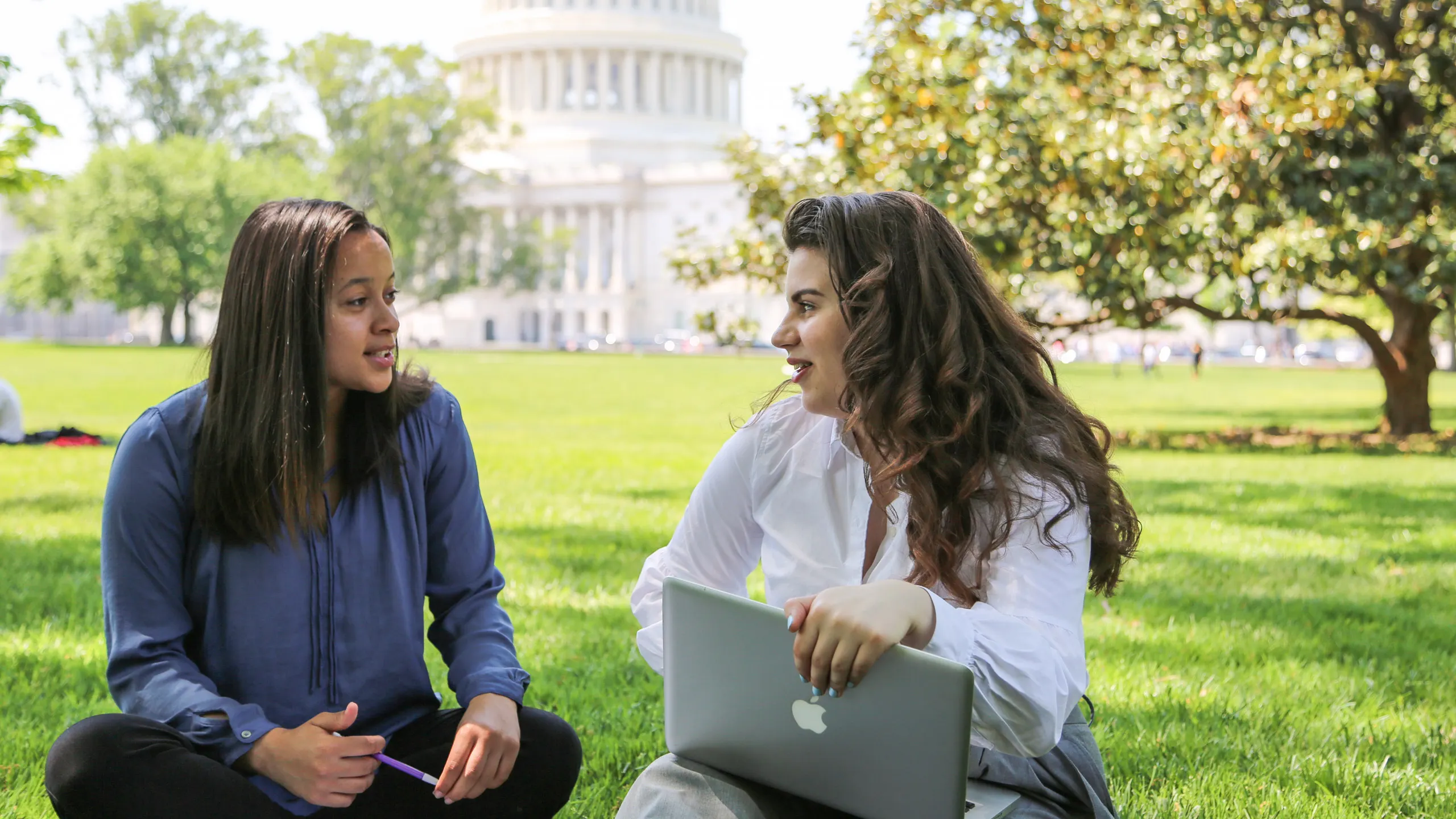 Two students sitting on the lawn talking, with computers in their laps, in front of the U.S. Capitol building