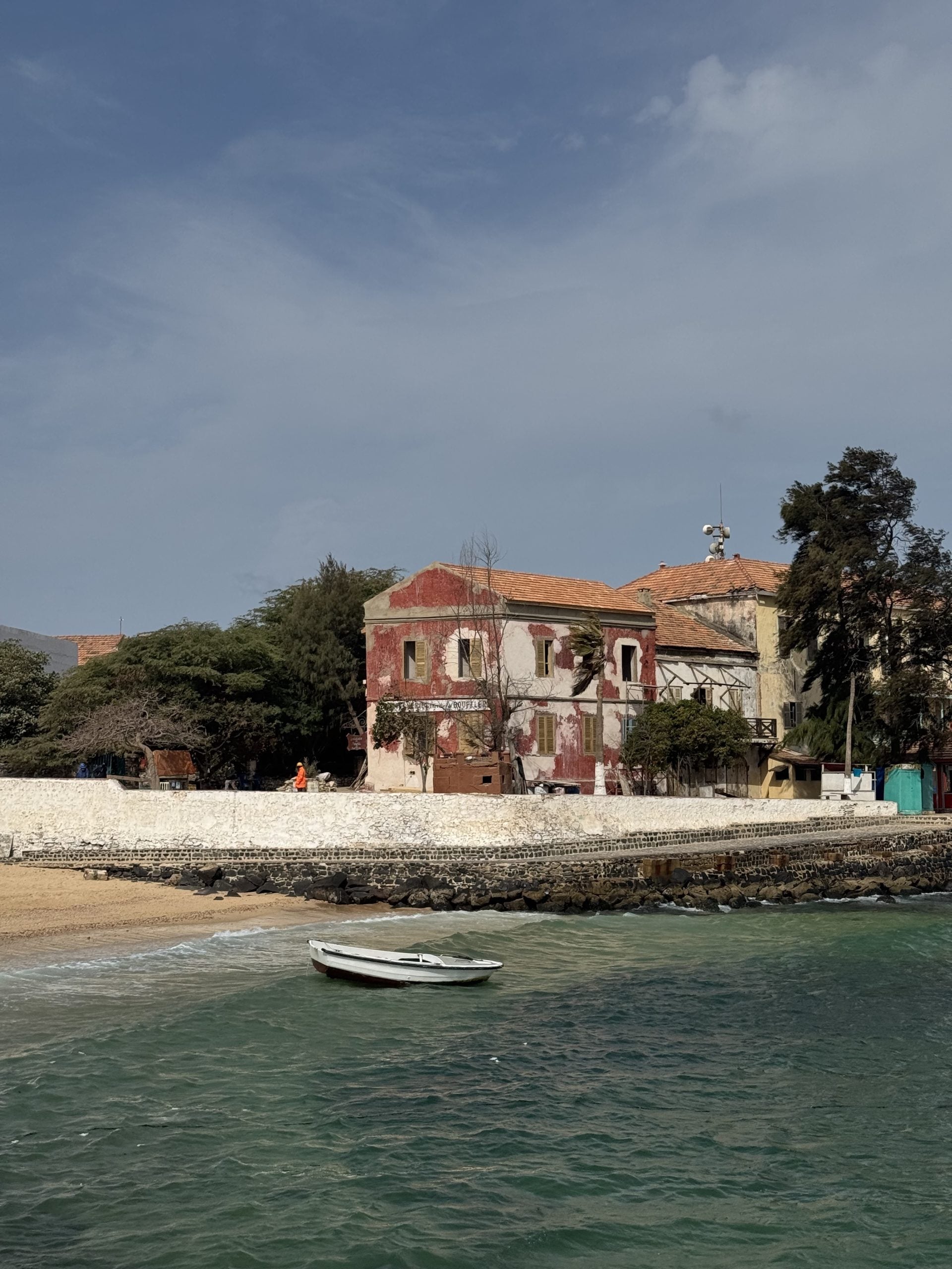 View of an old, weathered building with faded red walls and a terracotta roof next to the azure sea, under a clear blue sky, with a small boat floating nearby.