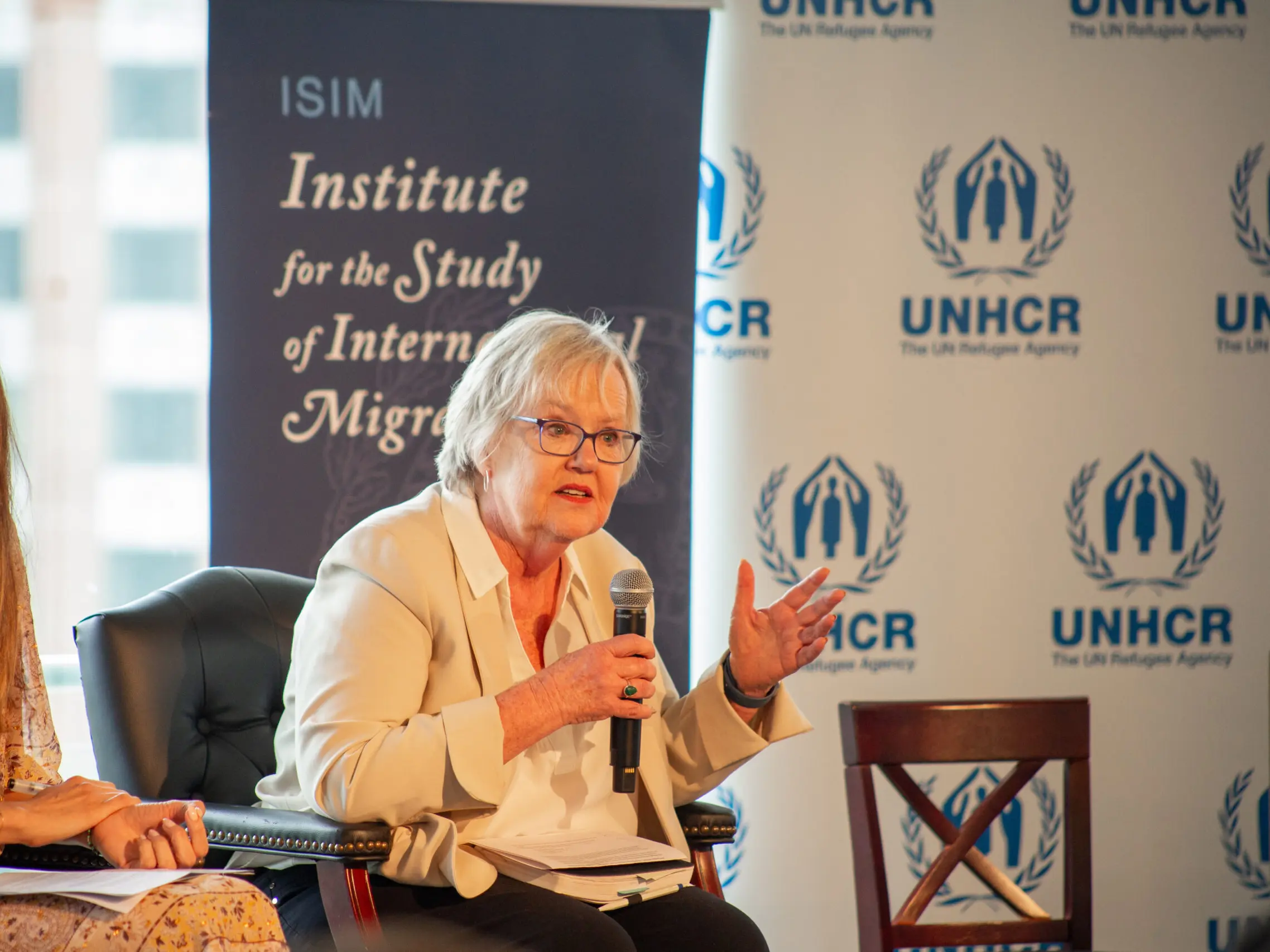 Professor Elizabeth Ferris speaking on stage with ISIM and UNHCR banners in the background