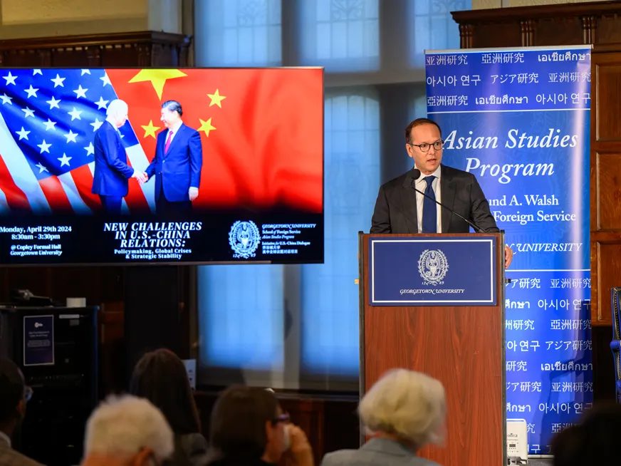 Prof. Medeiros speaking on a podium to a crowd. Behind him the banner for the Asian Studies Program. Next to him a screen the the USA and China flags.