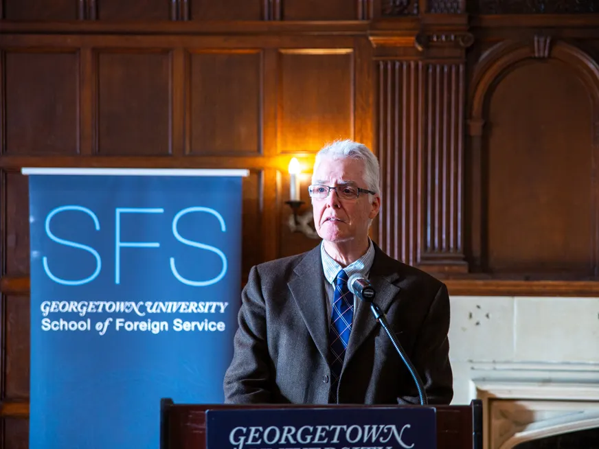 Alan Tidwell wearing a suit, speaking in front of a podium, behind him a banner for the School of Foreign Service