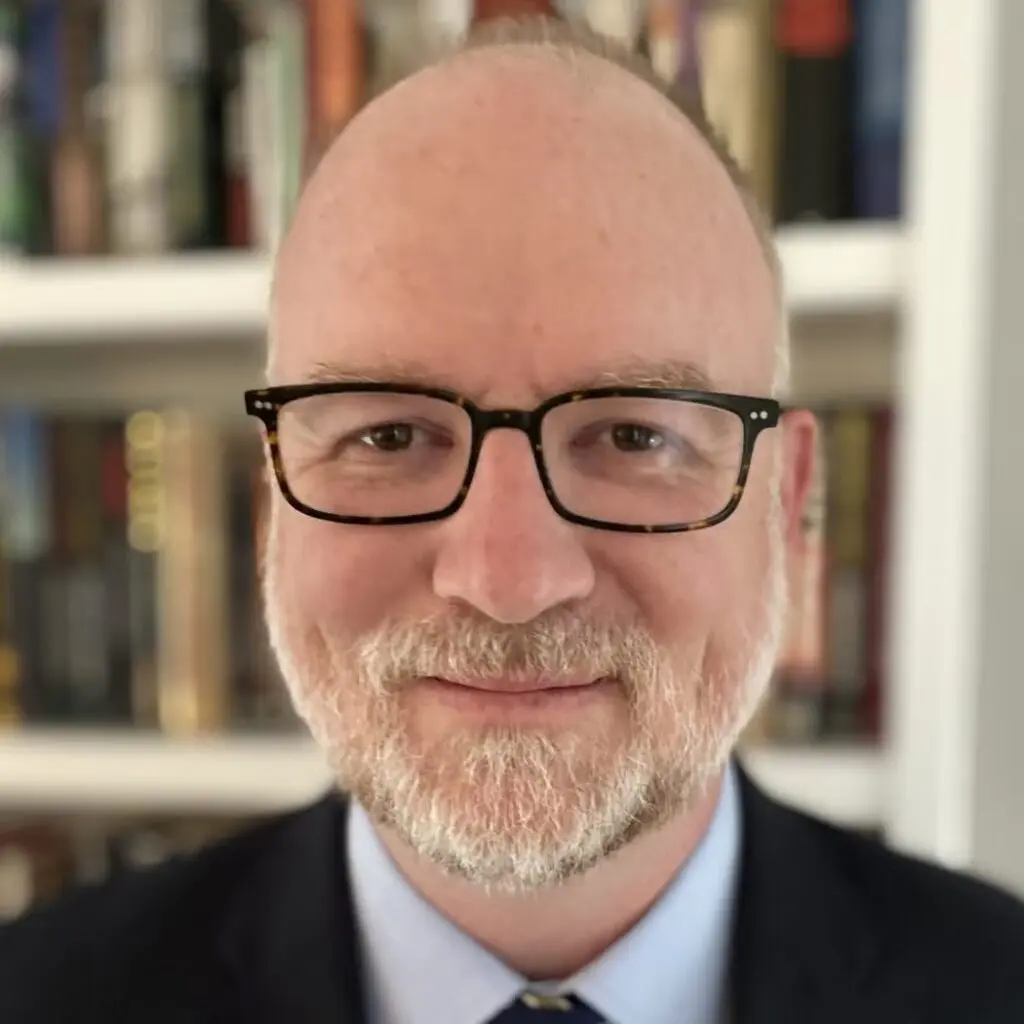 Andrew Amunson wearing a suit, tie, and black glasses, posing against a bookshelf.