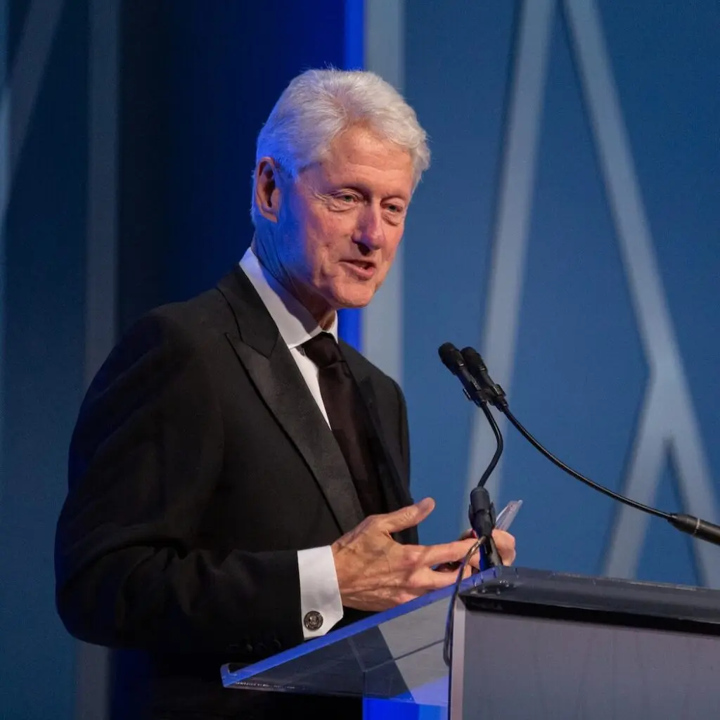 President Clinton standing at a podium in a black suit, giving a speech.