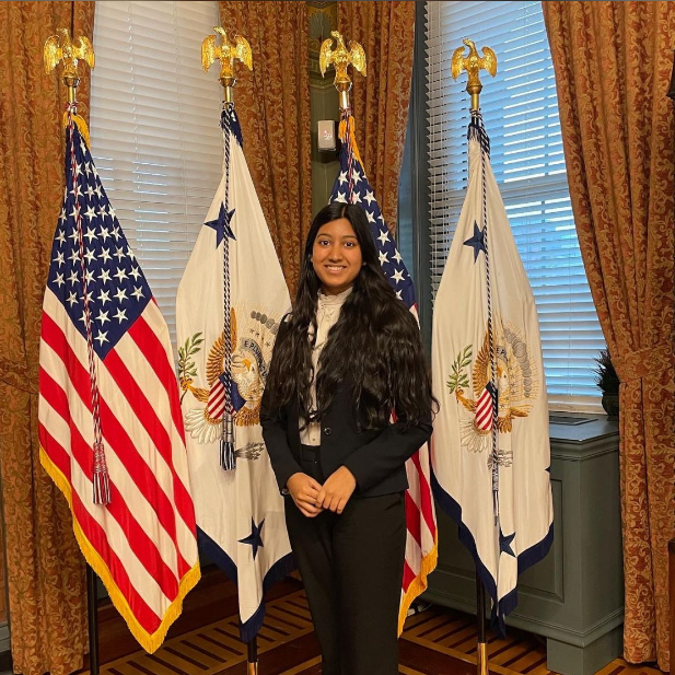 A person standing amongst U.S. flags and a flag with a blue and white emblem in a formal office setting.