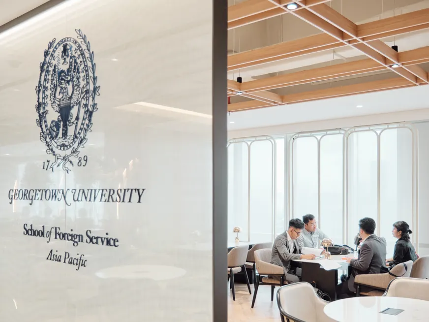 Four students sitting across from each other in a table, while speaking and browsing papers. In front of them a wall with the Georgetown University seal and the name School of Foreign Service Asia Pacific