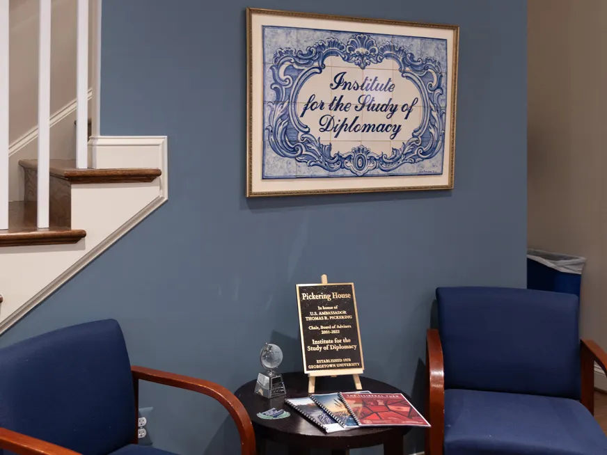 Indoors. A frame, with the words "Institute for the Study of Diplomacy" hangs on a blue wall. Underneath it, two blue chairs and a coffee table.