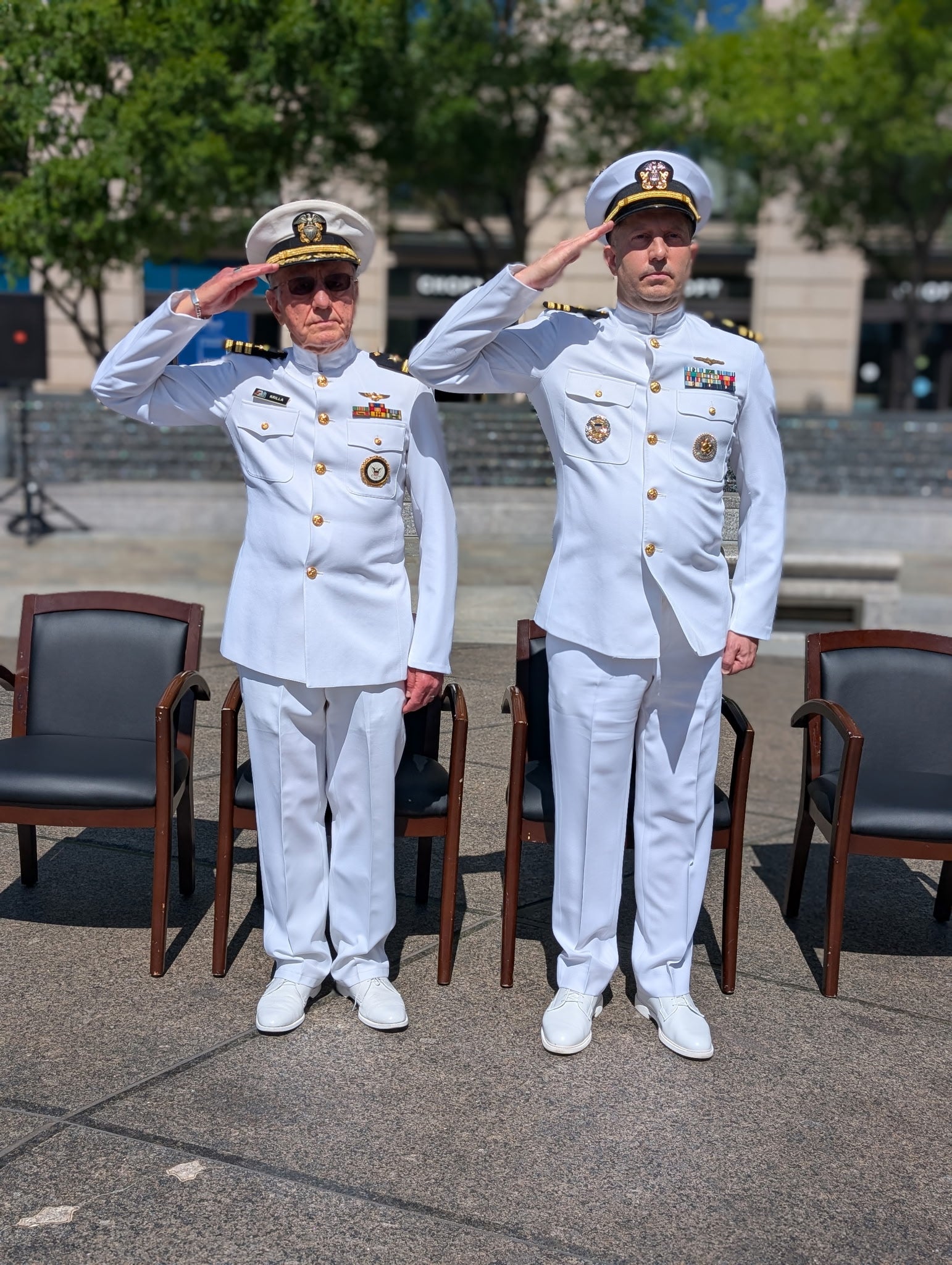 Two Navy officers in white uniforms saluting, standing outdoors.