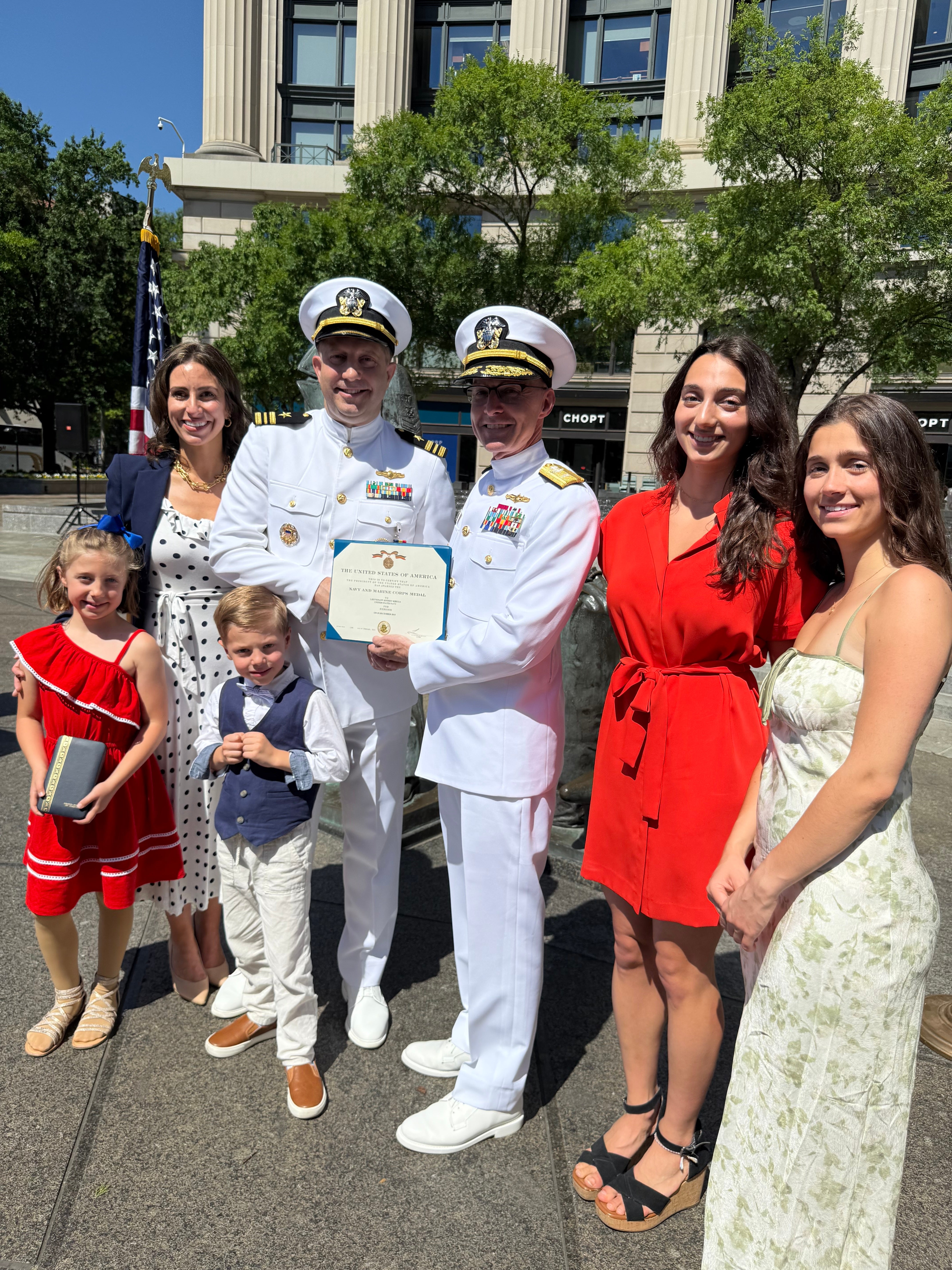 A group of seven people, including children, standing in front of a building on a sunny day during a formal military award ceremony.