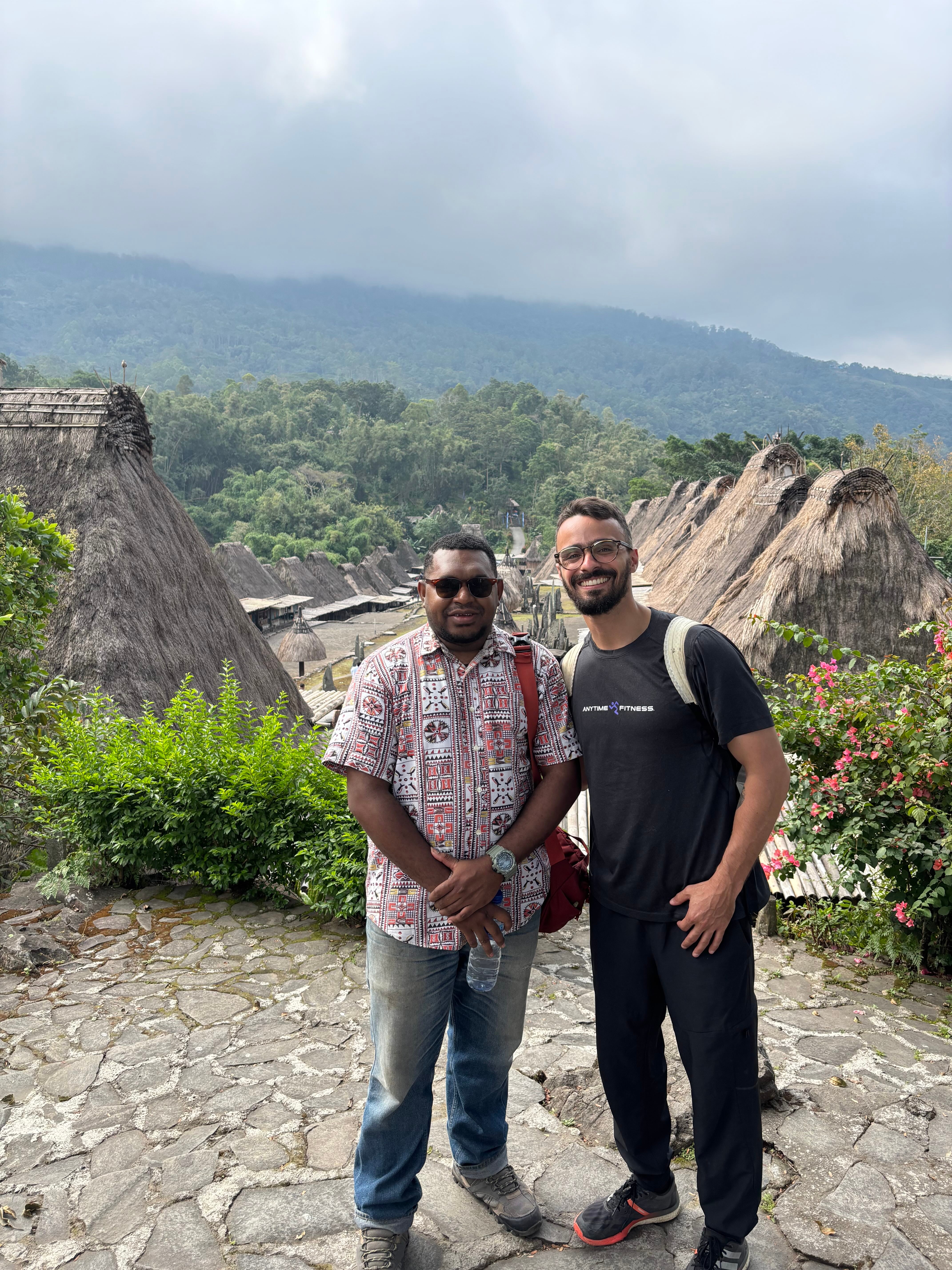Two men standing on a cobblestone path with traditional thatched-roof houses and a forested mountain in the background.