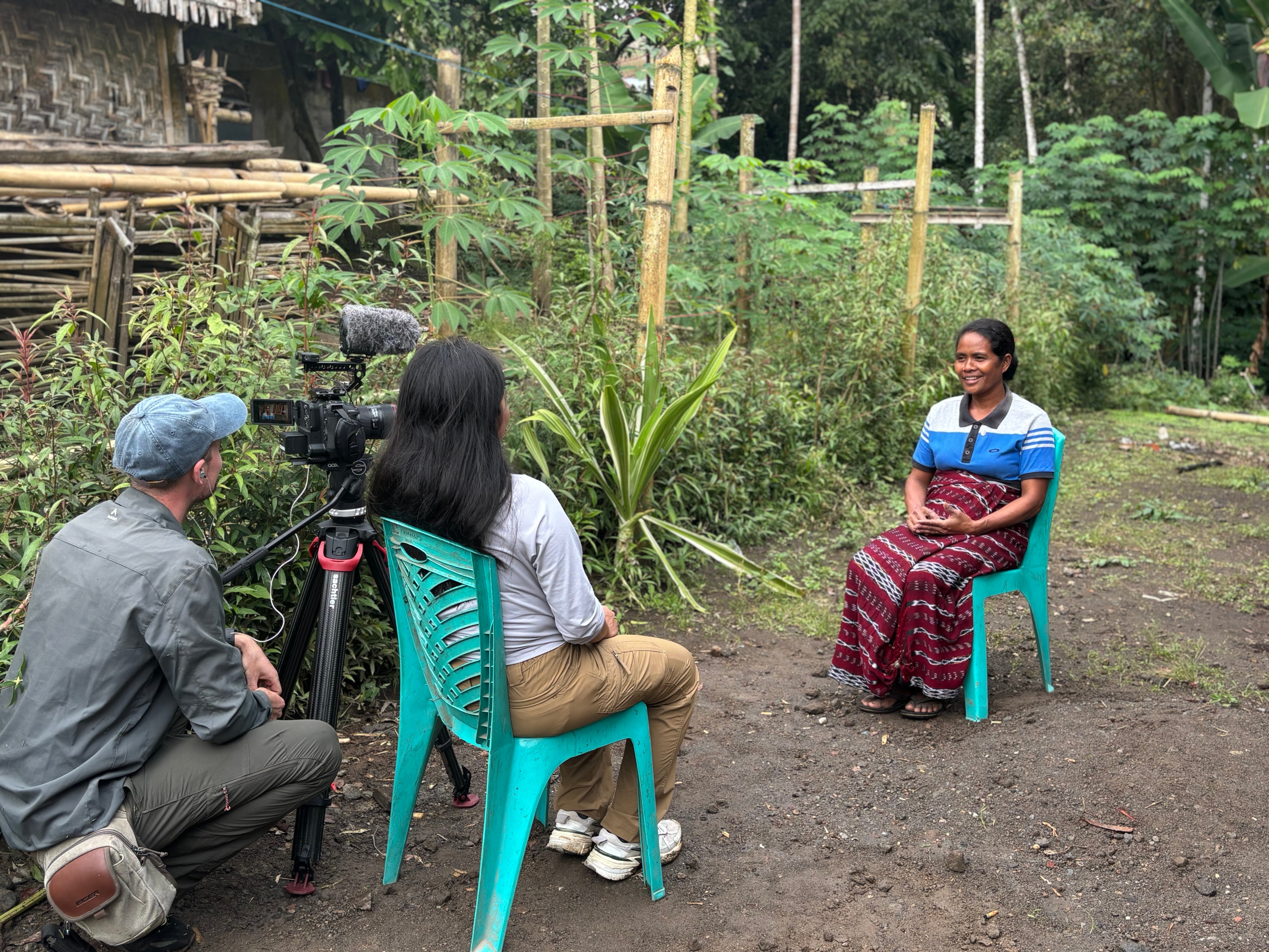 Two individuals, one operating a camera and the other holding a microphone, are conducting an interview with a seated person in traditional attire, in a rural setting with lush greenery and wooden structures visible in the background.