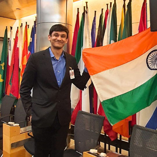 Person smiling and posing next to the Indian flag in a room with multiple international flags.