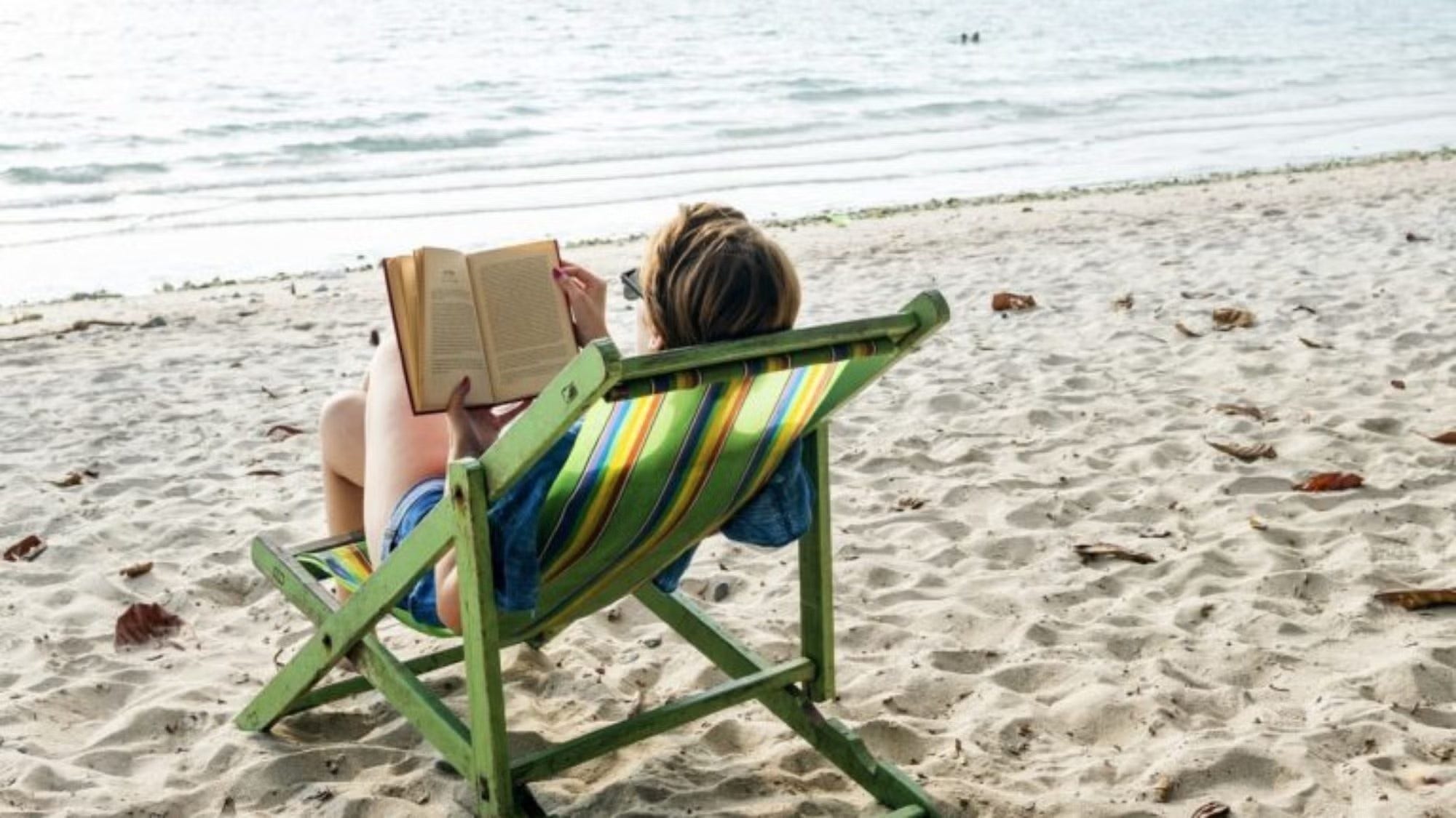 Woman in green and yellow striped reclining chair on beach, reading book with ocean in the background.