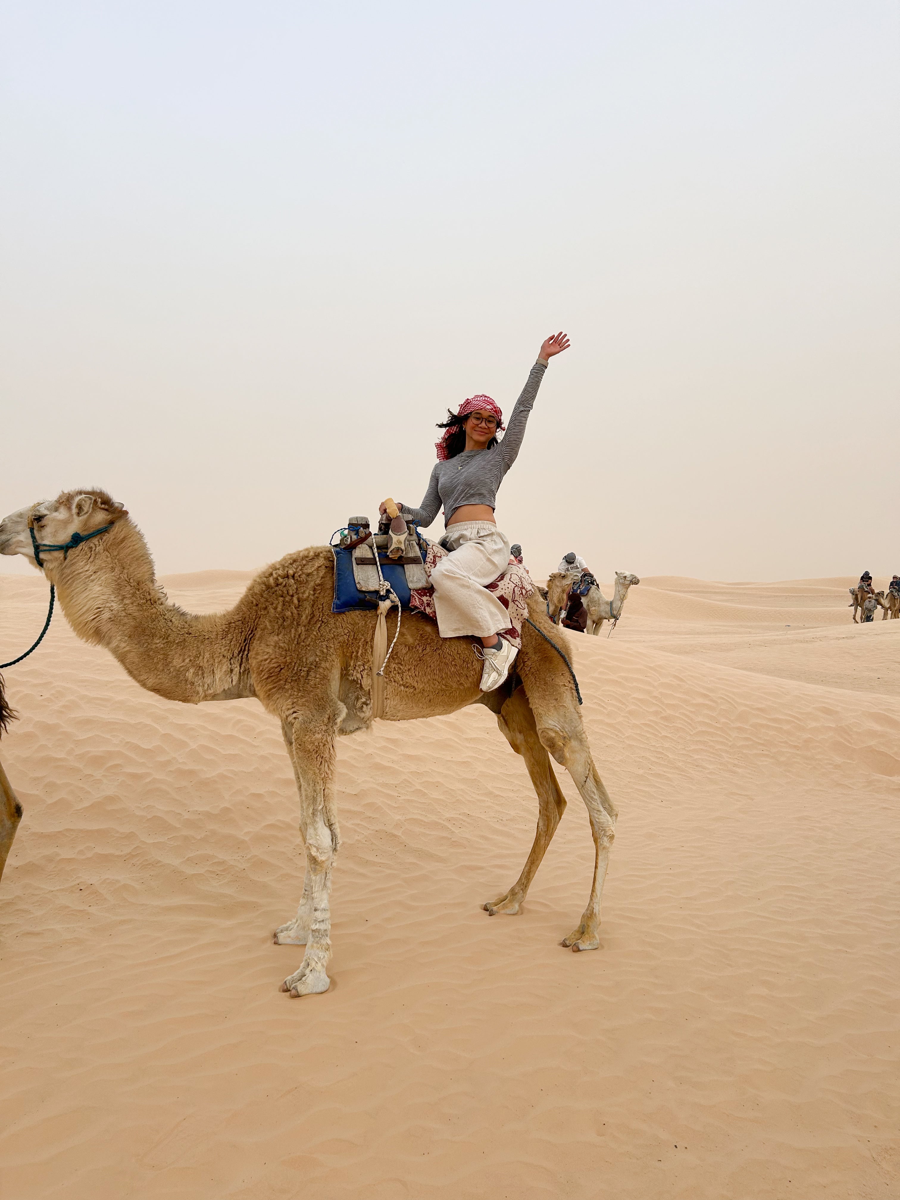 Woman riding a camel in the desert, waving cheerfully.