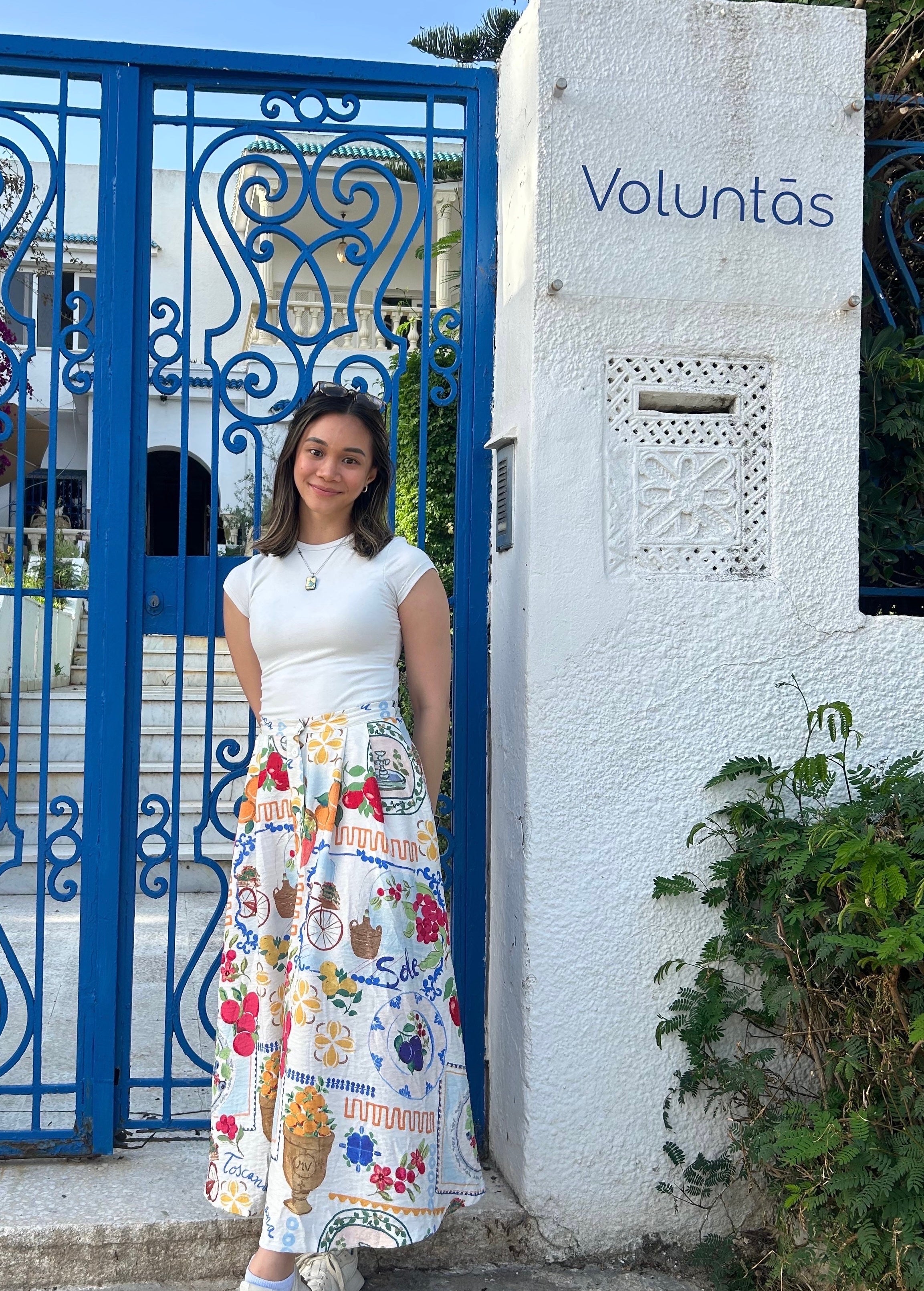 A woman wearing a long colorful skirt standing by a blue gate with the word "Voluntas" written on the adjacent white wall.