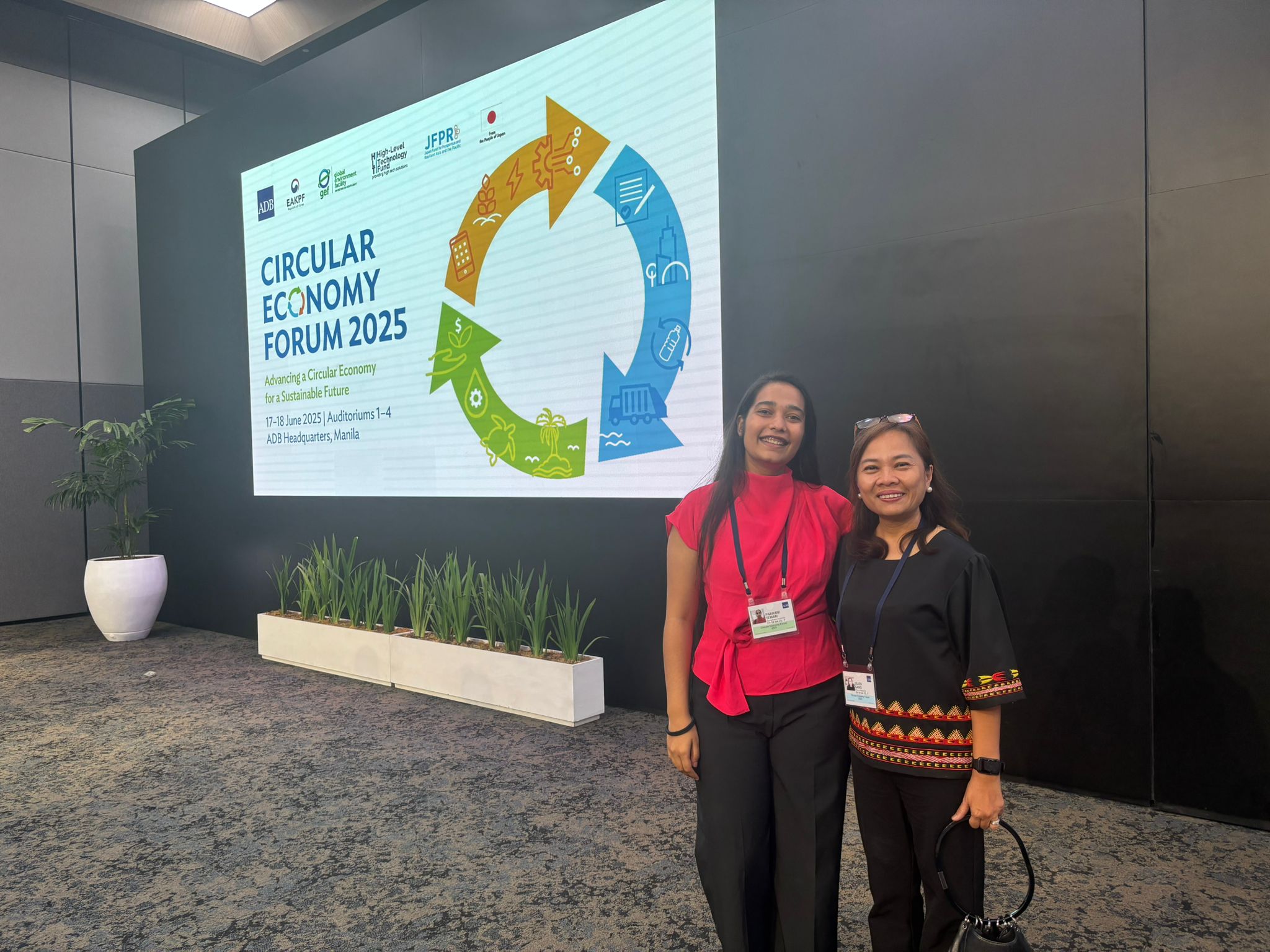 Two attendees smiling at a "Circular Economy Forum 2025" event, standing in front of a presentation screen displaying the forum's title and graphics about sustainability.
