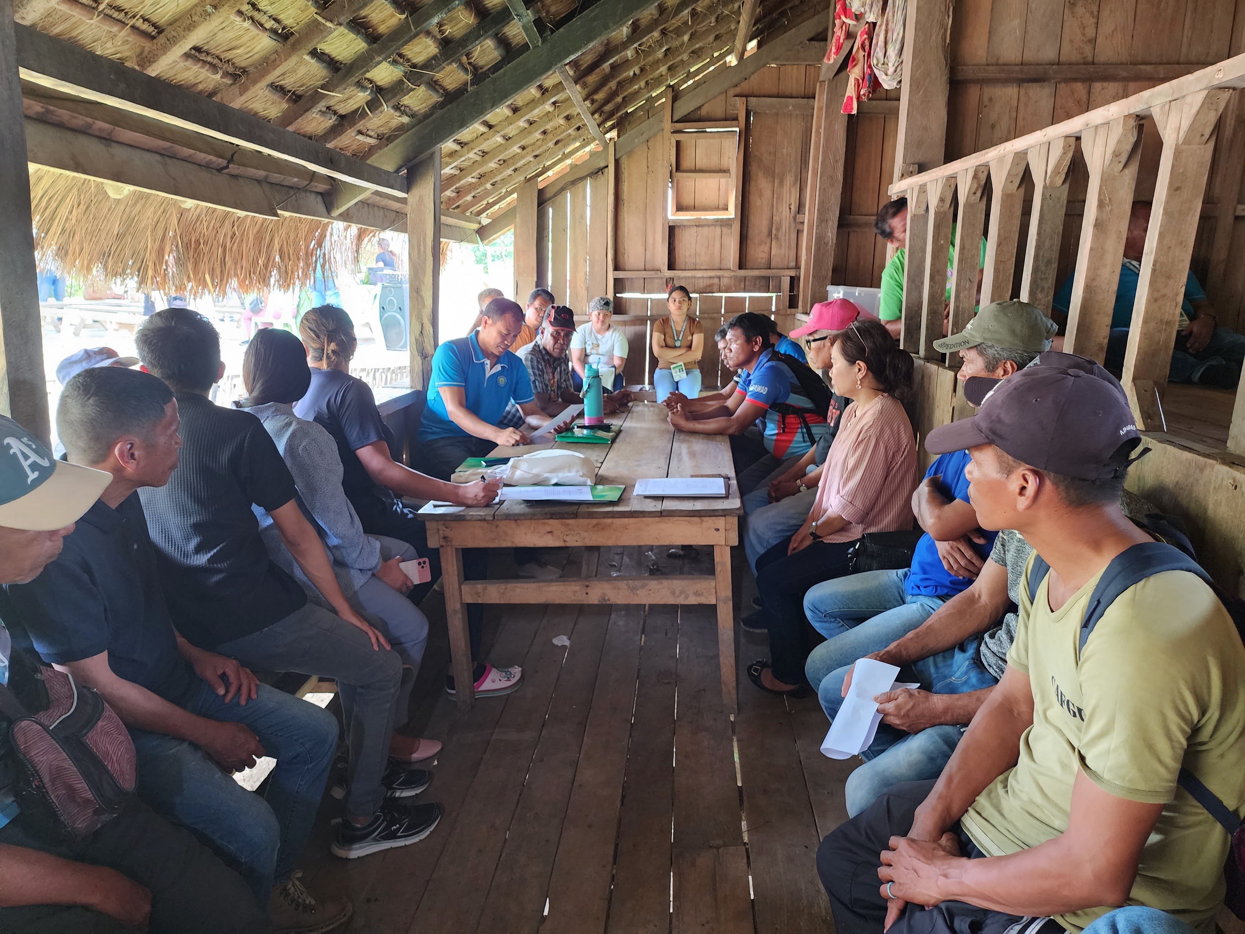 Group of people sitting around a long table in a wooden, open-sided structure, engaged in a meeting with papers and documents visible on the table.