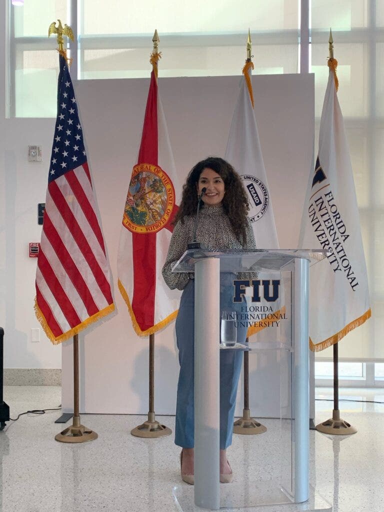 Photo of Pierina Valentina Anton Lopez speaking at a podium at Florida International University, in front of the U.S., Florida, USAID, and Florida International University flags.