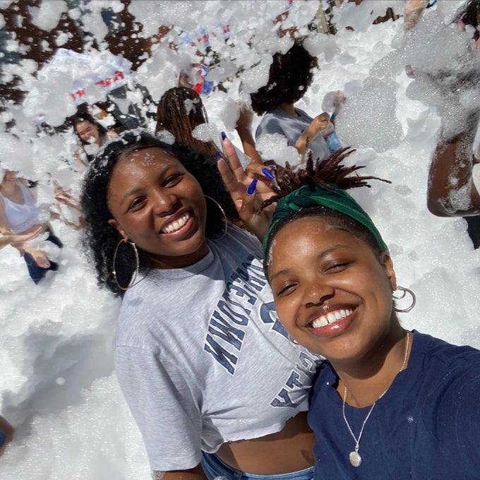Joella Kiondo smiling with another young woman, behind them smiling young people playing with foam bubbles.