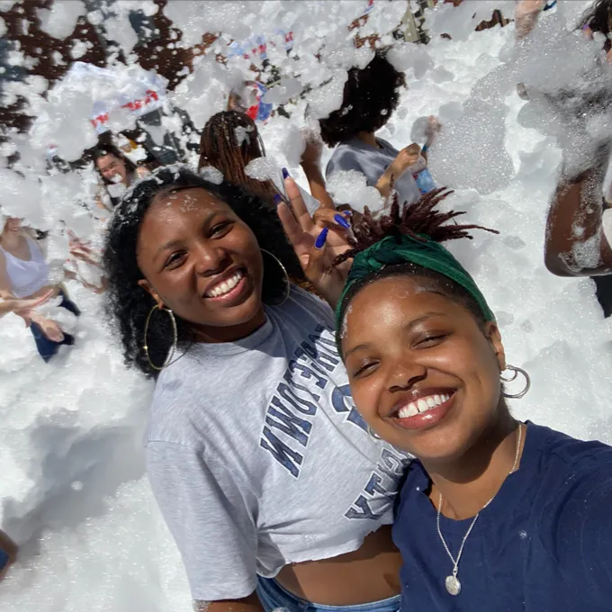 Joella Kiondo smiling with another young woman, behind them smiling young people playing with foam bubbles.