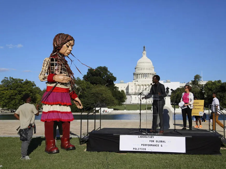 A man speaking on a stage in front of the US Capitol building. Next to him large 12 ft puppet of a woman. The stage says Laboratory for Global Performance and Politics