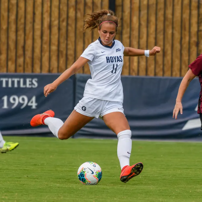 Maya Fernandez Powell in the soccer field, with a Hoyas #11 uniform on. She is kicking the ball in the middle of a run.